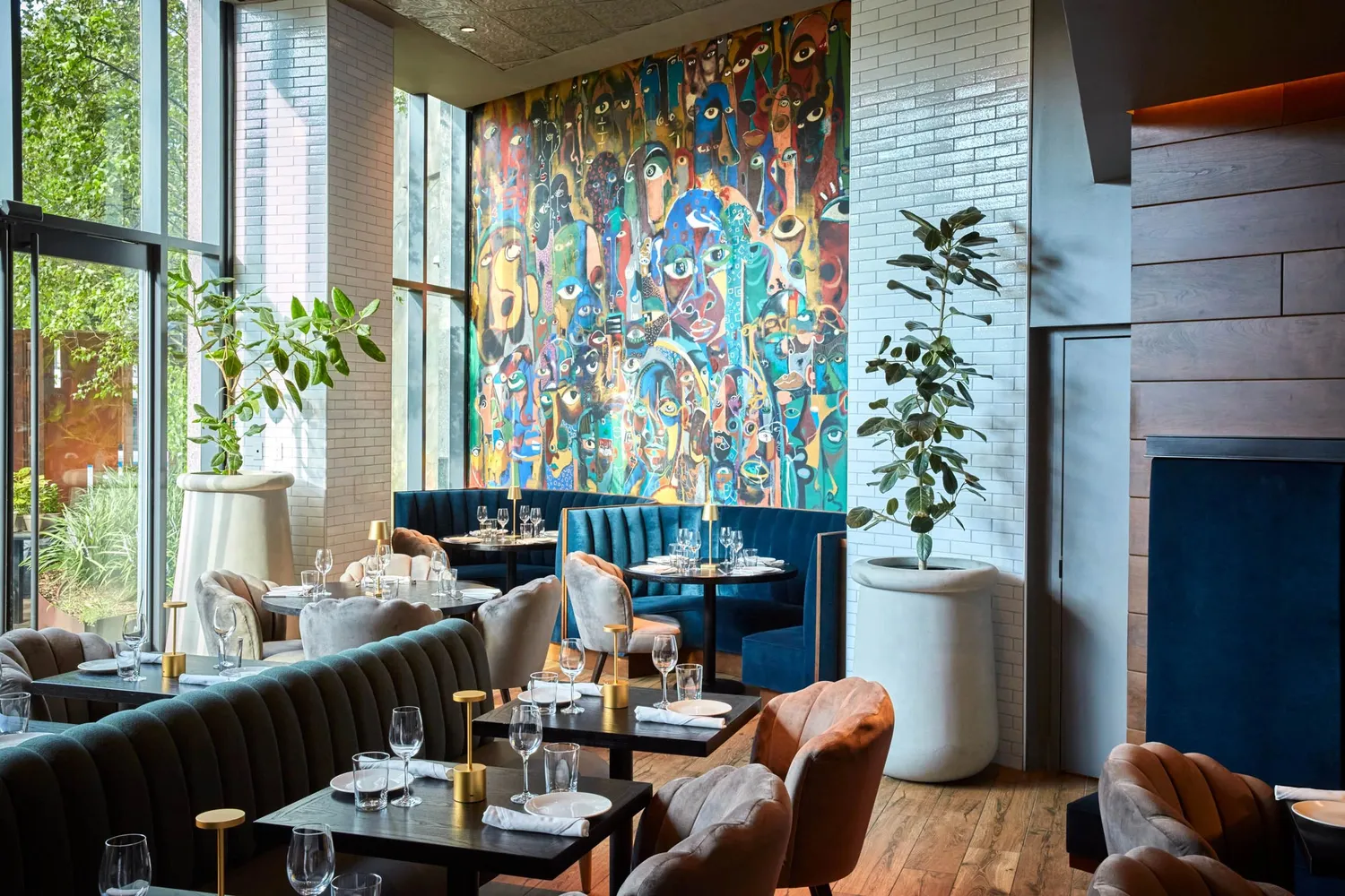 A modern restaurant interior with large windows, vibrant mural, and elegant table settings featuring dark velvet seating and tall plants in white pots.